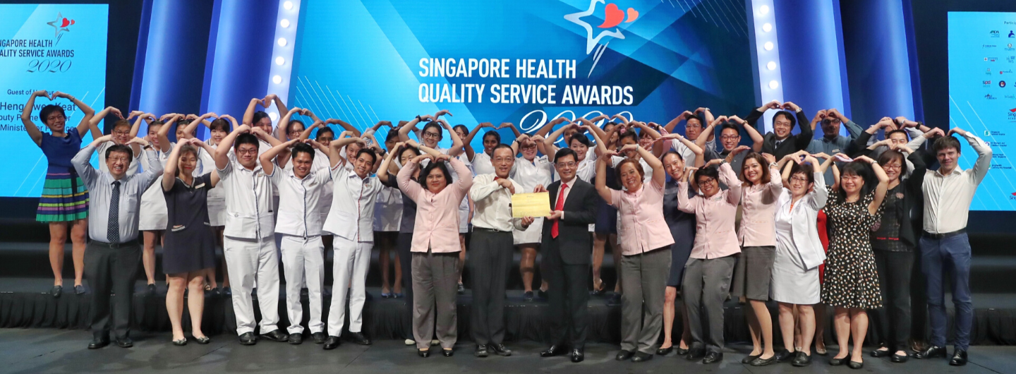 Group of people making heart shapes overhead on stage at Singapore Health Quality Service Awards.
